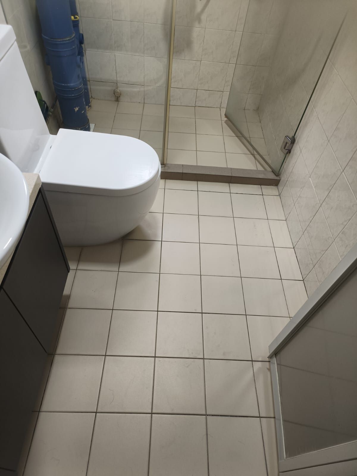 Before: Condo bathroom floor with old tiles, wall-hung toilet, and glass shower partition